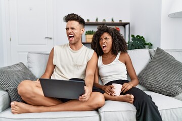 Fototapeta premium Young interracial couple using laptop at home sitting on the sofa angry and mad screaming frustrated and furious, shouting with anger. rage and aggressive concept.