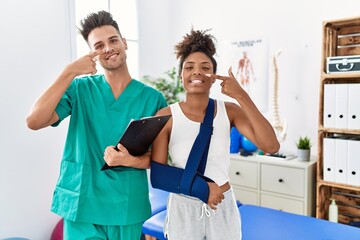 Fototapeta premium Physiotherapist working with patient wearing arm on sling at rehabilitation clinic pointing with hand finger to face and nose, smiling cheerful. beauty concept