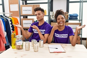 Young interracial people wearing volunteer t shirt at donations stand amazed and smiling to the camera while presenting with hand and pointing with finger.