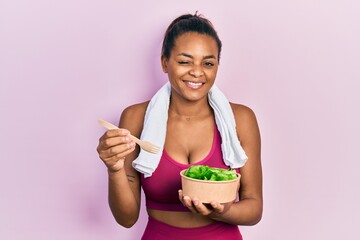 Young african american girl wearing sportswear eating salad winking looking at the camera with sexy expression, cheerful and happy face.