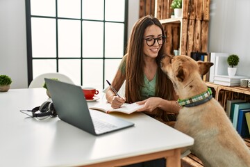 Young hispanic woman using laptop and writing on book sitting on table with dog at home