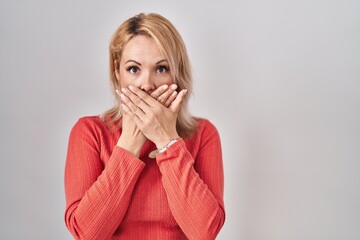 Blonde woman standing over isolated background shocked covering mouth with hands for mistake....