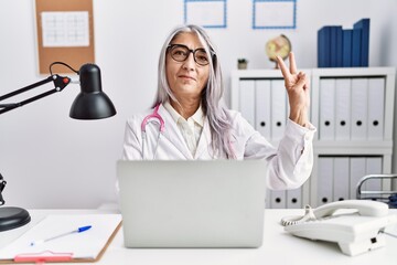 Middle age grey-haired woman wearing doctor uniform working using computer laptop smiling with happy face winking at the camera doing victory sign. number two.