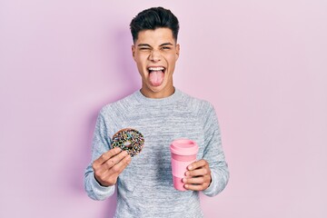 Young hispanic man eating doughnut and drinking coffee sticking tongue out happy with funny...