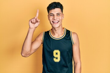 Young hispanic man wearing basketball uniform pointing finger up with successful idea. exited and...