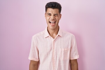 Young hispanic man standing over pink background sticking tongue out happy with funny expression. emotion concept.