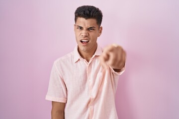 Young hispanic man standing over pink background pointing displeased and frustrated to the camera,...