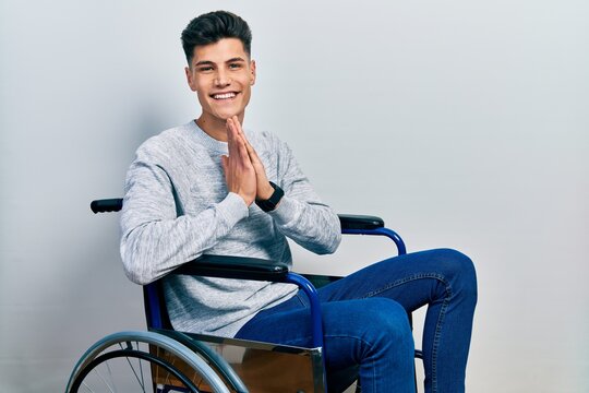 Young hispanic man sitting on wheelchair praying with hands together asking for forgiveness smiling confident.