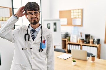 Hispanic man with beard wearing doctor uniform and stethoscope at the office confuse and wonder about question. uncertain with doubt, thinking with hand on head. pensive concept.