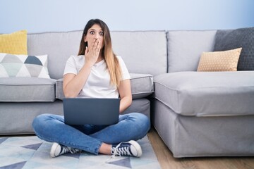 Obraz premium Young brunette woman sitting on the floor at home using laptop covering mouth with hand, shocked and afraid for mistake. surprised expression