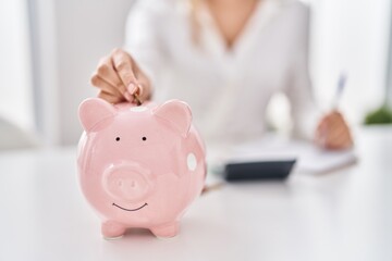 Young woman inserting coin on piggy bank at home - obrazy, fototapety, plakaty