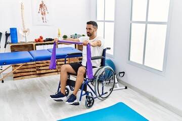 Young hispanic man patient having rehab session using elastic band sitting on wheelchair at clinic