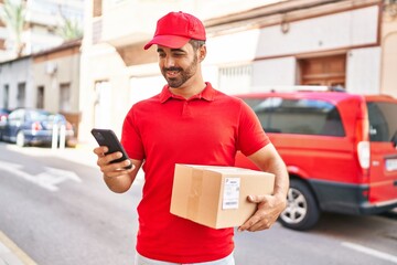 Young hispanic man courier using smartphone holding package at street - obrazy, fototapety, plakaty