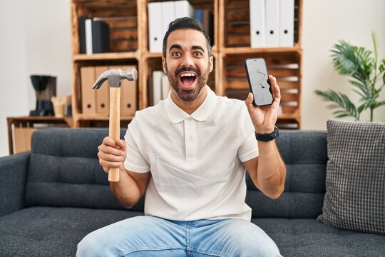 Young hispanic man with beard holding hammer and broken smartphone showing cracked screen celebrating crazy and amazed for success with open eyes screaming excited.
