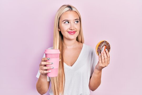 Young blonde girl eating doughnut and drinking coffee smiling looking to the side and staring away thinking.