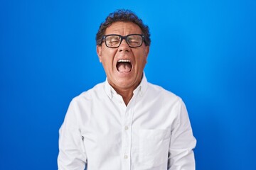 Middle age hispanic man standing over blue background angry and mad screaming frustrated and furious, shouting with anger. rage and aggressive concept.