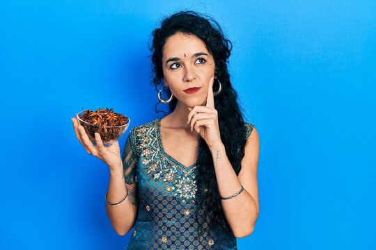 Young woman wearing bindi and traditional kurta dress holding bowl of star anise serious face thinking about question with hand on chin, thoughtful about confusing idea