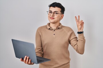 Non binary person using computer laptop smiling looking to the camera showing fingers doing victory...