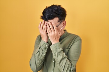 Non binary person standing over yellow background with sad expression covering face with hands while crying. depression concept.