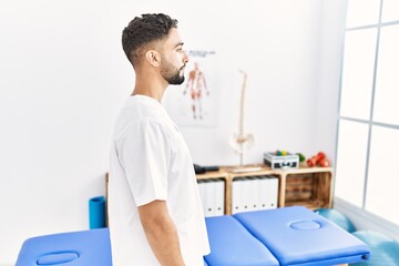 Young handsome man with beard working at pain recovery clinic looking to side, relax profile pose with natural face with confident smile.