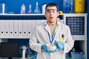 Young arab man working at scientist laboratory in shock face, looking skeptical and sarcastic, surprised with open mouth