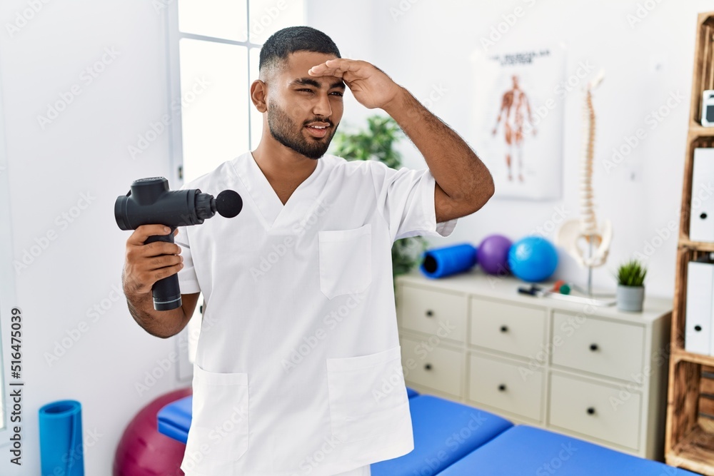 Sticker Young indian physiotherapist holding therapy massage gun at wellness center very happy and smiling looking far away with hand over head. searching concept.