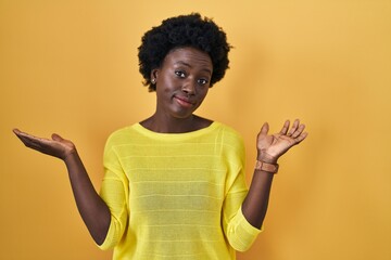 African young woman standing over yellow studio clueless and confused expression with arms and hands raised. doubt concept.