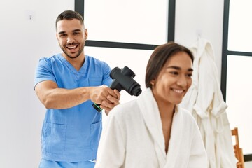 Obraz premium Latin man and woman wearing physiotherapy uniform massaging neck using percussion gun at beauty center