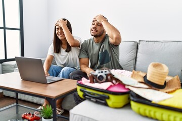 Young interracial couple packing summer clothes in suitcase looking at laptop stressed and frustrated with hand on head, surprised and angry face