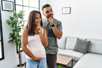 Young interracial couple expecting a baby, touching pregnant belly with hand on chin thinking about...