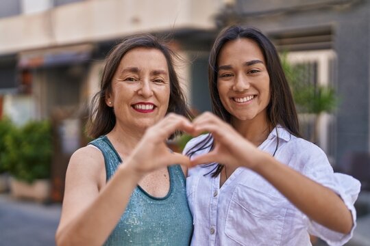 Two women mother and daughter smiling confident doing heart gesture with hands at street