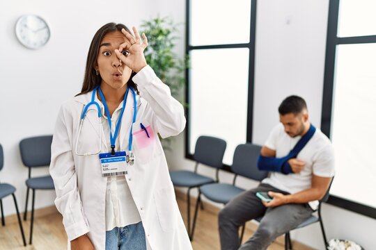 Young asian doctor woman at waiting room with a man with a broken arm doing ok gesture shocked with surprised face, eye looking through fingers. unbelieving expression.