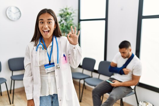 Young asian doctor woman at waiting room with a man with a broken arm smiling positive doing ok sign with hand and fingers. successful expression.
