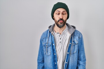 Young hispanic man with tattoos wearing wool cap making fish face with lips, crazy and comical gesture. funny expression.