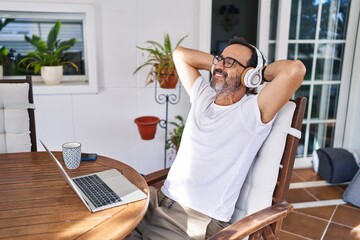 Middle age man relaxed with hands on head using laptop and headphones at terrace home