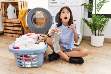 Young hispanic girl doing laundry holding socks amazed and surprised looking up and pointing with fingers and raised arms.