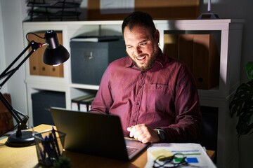 Plus size hispanic man with beard working at the office at night winking looking at the camera with sexy expression, cheerful and happy face.