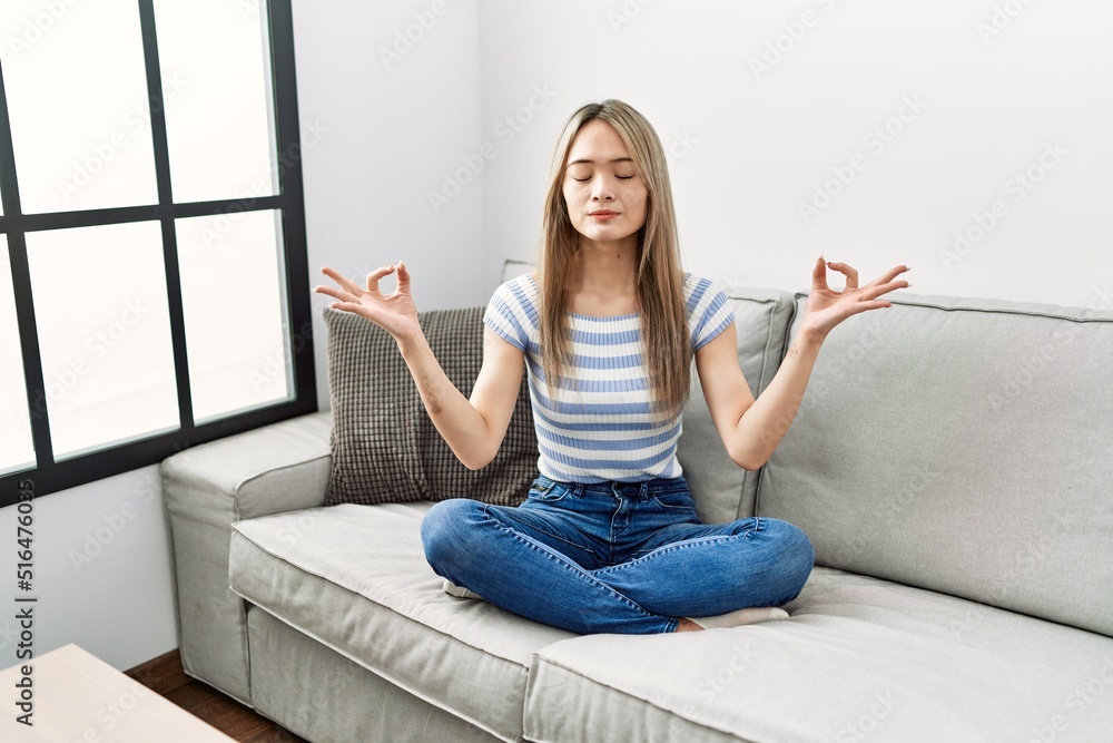 Sticker asian young woman sitting on the sofa at home relax and smiling with eyes closed doing meditation ge