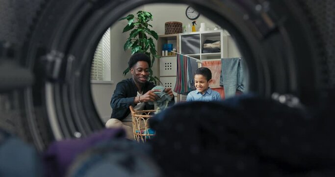 A view from inside the drum of a young man and his son having fun doing housework, throwing more clothes into the washing machine, and competing to see who can hit it more times.