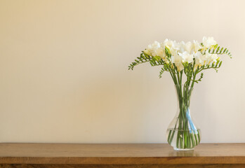 Closeup of white freesia flowers in glass jar on oak side table against beige wall with copy space (selective focus)