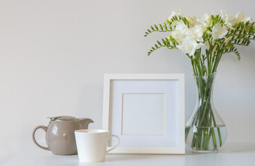White freesia flowers in glass vase with blank square picture frame, tea cup and teapot against white background (selective focus)