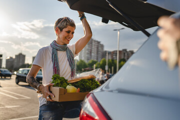 One woman mature caucasian female standing by the back trunk of her car on the parking lot of the supermarket shopping mall or grocery store with vegetables food in box putting them in the vehicle - obrazy, fototapety, plakaty