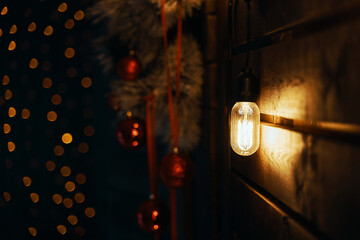 Christmas decorations. Vintage type bulb and fir branches with red balls and ribbons on wooden wall. Copy space for New Year congratulations. Blurred warm garland lights on background. Party mood.