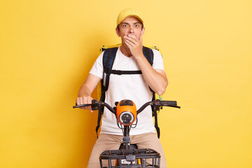 Horizontal shot of shocked deliveryman on bicycle wearing white T-shirt and cap isolated over...