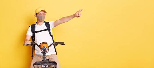 Indoor shot of young adult Caucasian delivery man on bycycle wearing white t shirt, cap and protective mask, delivering orders during quarantine, pointing aside, showing copy space for advertisement.