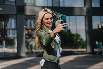 Young hipster Italian girl making selfie touching blonde hair against modern building. Beautiful...