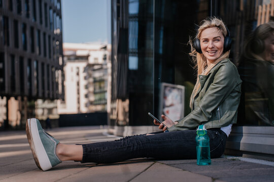 Cheerful young blonde woman in headphones wearing casual clothes sitting on the ground with water bottle happy listening music on weekend. Cheerful italian hipster girl enjoying life and youth.