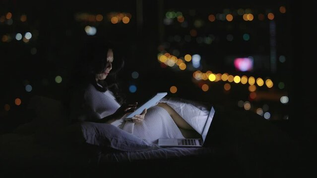 Woman using wireless internet connection on laptop and tablet, planning, booking hotel for travel, booking flight tickets, sitting on bed in a dark room. Blurred bokeh background, city lights at night