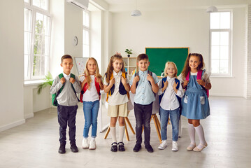 Portrait of small smiling group of first graders in modern elementary school classroom. Little boys and girls in different clothes and with backpacks on their shoulders stand in row and look at camera