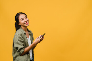 Surprised young Asia lady using mobile phone with positive expression, smile broadly, dressed in casual clothing and looking at camera on yellow background. Happy adorable glad woman rejoices success.